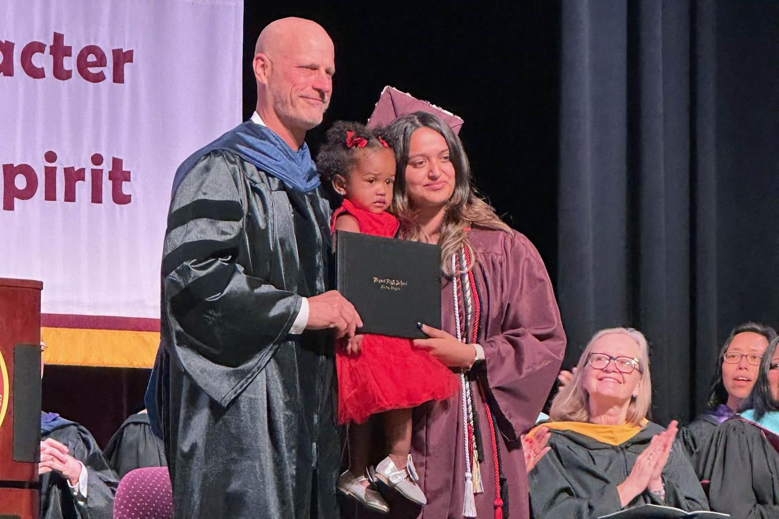 Newly-minted Bryant High School graduate Anyeli Salguero receives her diploma from principal Chris Larrick with her 20-month-old daughter in her arms. School board member Karen Corbett Sanders claps after listening to Anyeli's speech.